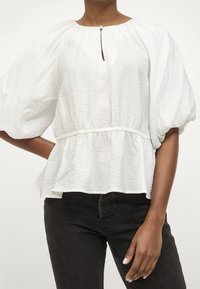 Blouse blanche texturée avec un col rond, détail en trou de serrure et taille froncée. Présente des manches bouffantes et un ourlet peplum. Associée à un pantalon noir.