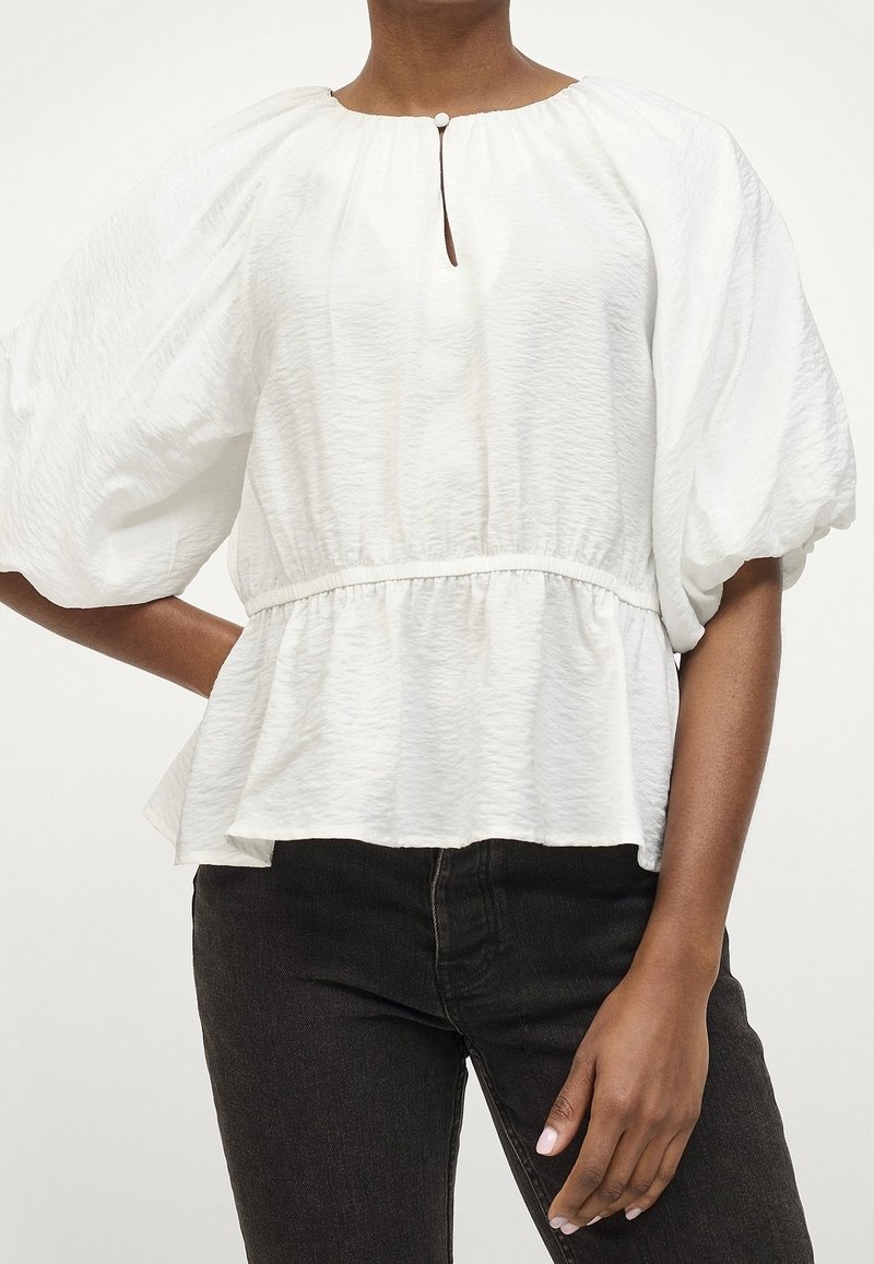 Blouse blanche texturée avec un col rond, détail en trou de serrure et taille froncée. Présente des manches bouffantes et un ourlet peplum. Associée à un pantalon noir.