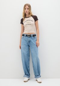 Mujer joven con cabello largo rubio que lleva una camiseta recortada beige y marrón de "Club Retriever", jeans azules sueltos, cinturón negro y zapatillas blancas.