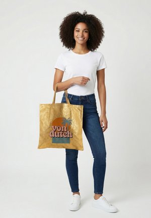 Mujer sonriente con cabello rizado que lleva una camiseta blanca y vaqueros, sosteniendo un bolso tote dorado con un sol naranja, una palmera y el texto "von dutch".