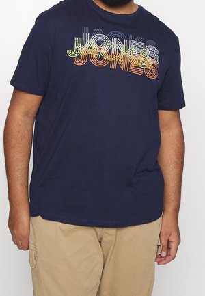 Mężczyzna ubrany w granatowy T-shirt z wielokolorowym, warstwowym napisem "JONES" oraz beżowe krótkie spodnie typu cargo, stojący na jednolitym tle.