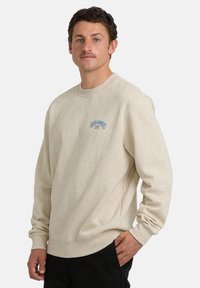 Mężczyzna z krótkimi kręconymi włosami i wąsami, ubrany w beżowy sweter z małym niebiesko-czerwonym logo, stoi z jedną ręką w kieszeni.