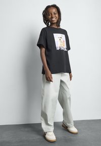 Smilende barn med dreadlocks som har på seg svart T-skjorte med hund og teksten "Sip Thea", lyse bukser og hvite sneakers, står på grått gulv.