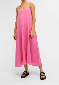 Robe maxi rose sans manches avec un col en V, fabriquée en tissu texturé. Dotée de fines bretelles et d'une coupe ample. Portée avec des sandales noires.