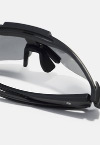Lunettes de soleil avec une monture noire élégante, des branches courbées et des verres teintés foncés. Elles disposent de coussinets de nez réglables et d'un logo discret sur le côté.