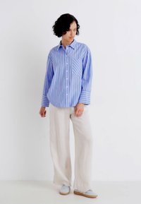 Camicia a righe blu e bianche con taschino sul petto, abbinata a pantaloni di lino larghi e chiari e scarpe azzurre chiare.