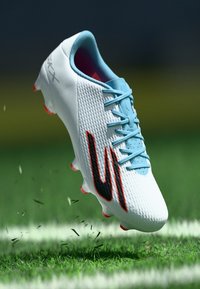 Scarpa da calcio bianca con accenti blu, tomaia texturizzata, logo nero e suola rossa, sospesa sopra l'erba con lievi detriti sottostanti.