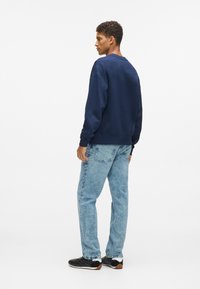 Jūrų mėlynos spalvos džemperis su apvalia iškirpte, derinamas su šviesiai mėlynomis džinsomis. Medžiaga atrodo minkšta, su laisvu siluetu ir minimaliais akcentais.