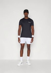 Μαύρο αθλητικό T-shirt με μικρό λογότυπο, συνδυασμένο με λευκό σορτς. Λευκά αθλητικά παπούτσια και βραχιολάκια στον καρπό. Το μοντέλο στέκεται όρθιο μπροστά σε ουδέτερο φόντο.