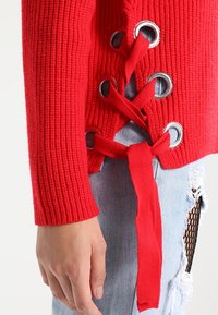 Red ribbed sweater with metallic grommets and red ties; fitted sleeves and side lace-up design. Worn with ripped light blue jeans.