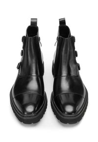 Coppia di stivaletti alla caviglia in pelle nera con suole spesse, cerniere laterali e tre fibbie decorative su ciascuno stivale.