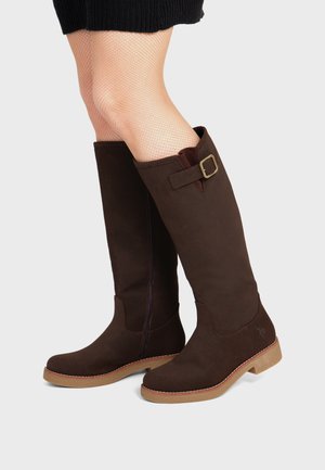 Jambes portant des bottes marron montantes avec détail de boucle et des bas résille, sur un fond clair uni.