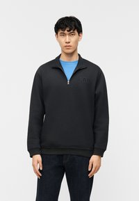 Νεαρός με κοντά μαύρα μαλλιά, ντυμένος με μαύρο φούτερ GAP με φερμουάρ πάνω από μπλε πουκάμισο και σκούρο τζιν, στέκεται μπροστά σε απλό ανοιχτό φόντο.