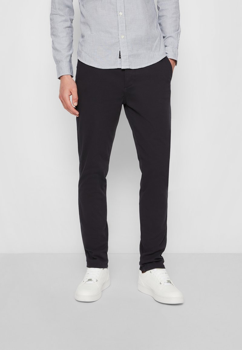 Homme portant un pantalon ajusté noir, des baskets blanches et une chemise rayée bleu clair à boutons, debout avec une main dans la poche.