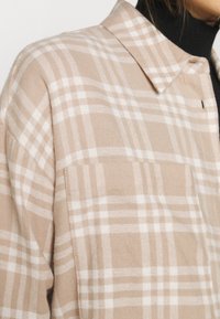 Veste-chemise à carreaux beige et blanche portée sur un col roulé noir, montrant le col et la partie supérieure de la poitrine.