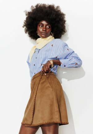 Femme aux grands cheveux bouclés portant une chemise rayée bleue, une écharpe claire, une jupe en daim marron et des bracelets en or, posant devant un fond blanc.