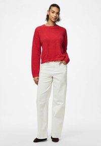 Maglione rosso a maglia con ampie maniche e orlo scallop, abbinato a pantaloni bianchi a gamba larga. L'outfit è completato da scarpe marroni scure.
