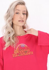 Rózsaszín, túlméretezett pulóver "Feelin' Blossom" grafikai mintával, amely virágokat és egy szivárványt ábrázol, sima anyagból, széles ujjal.