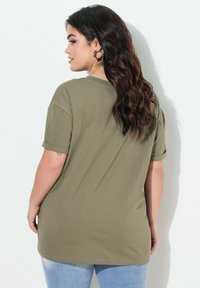 T-shirt oversize vert olive avec des manches courtes roulées, fabriqué en coton doux. Présenté de dos, montrant un ourlet droit et une coupe décontractée.