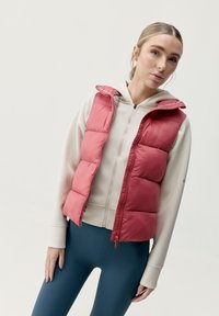 Magenta vadderad väst med huva, med dragkedjestängning och sydda paneler. I kombination med en ljusbeige zip-up hoodie och teal leggings.