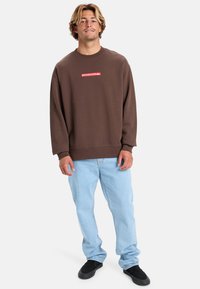 Bruine sweatshirt met een ronde hals en een rood "QUIKSILVER" logo. Gestyled met lichtblauwe spijkerbroek en zwarte schoenen. Zachte stof, losse pasvorm.