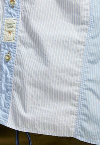 Camicia a righe blu chiaro con motivi verticali, chiusura con bottoni e etichetta del marchio. Tessuto morbido e texture liscia. Design a maniche corte.