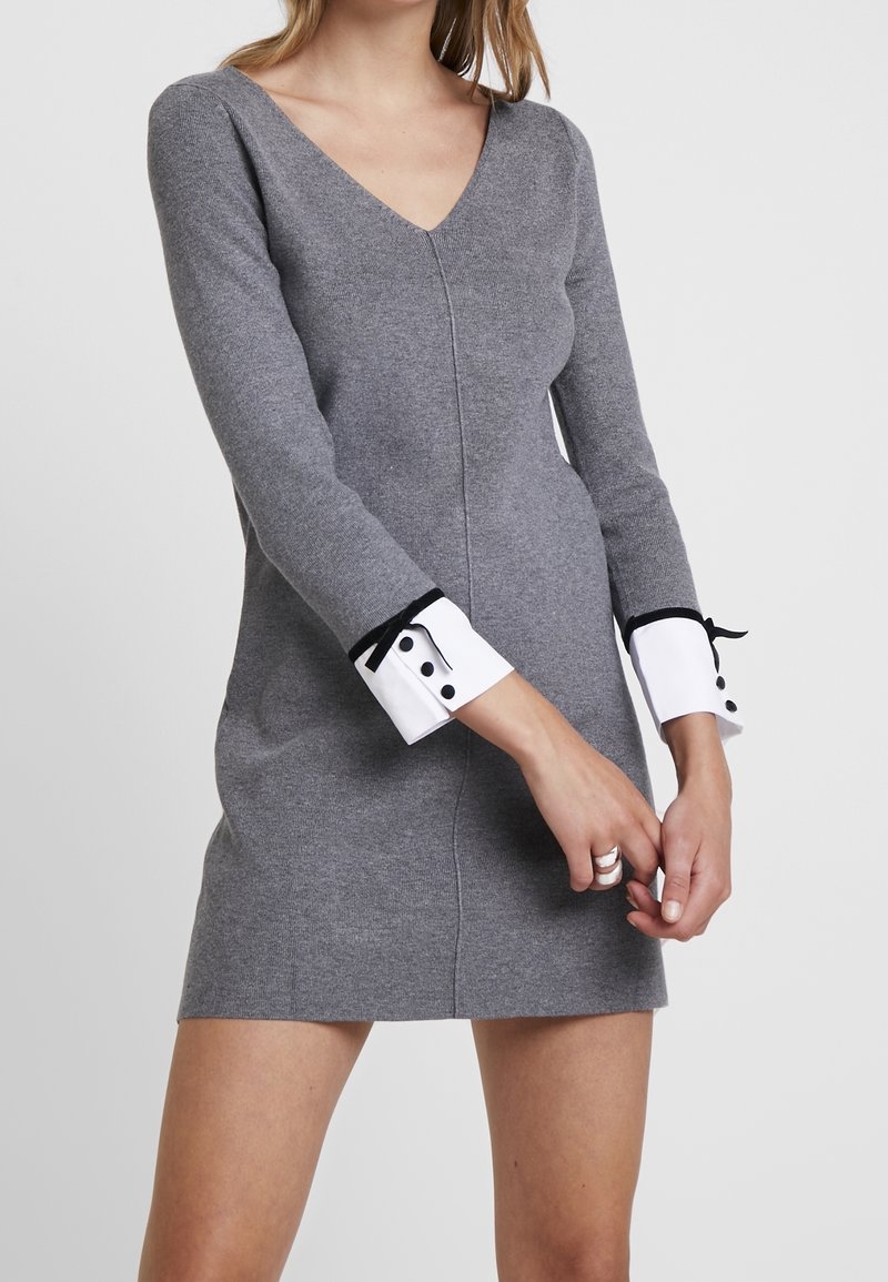 Robe tricotée grise avec un col en V, des manches longues et des poignets blancs ornés d'accents noirs et de boutons. Texture lisse, design ajusté.