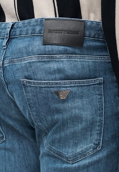 Modre kavbojke iz jeansa z usnjenim trakcem, na katerem je napis "EMPORIO ARMANI", in srebrnim logotipom orla na zadnjem žepu. Imajo izpiran učinek.