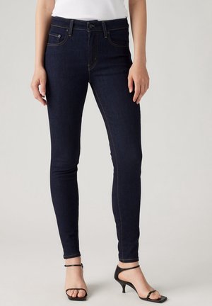 Femme portant un jean skinny bleu foncé et des talons ouverts noirs sur un fond clair uni.