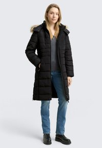 Zwarte pufferjas met een capuchon van bont, lange mouwen en horizontale quilting. Gecombineerd met een grijze top, blauwe jeans en zwarte laarzen.