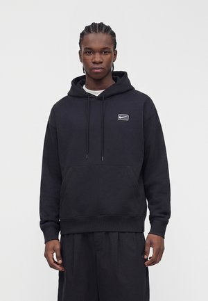 Młody mężczyzna z zaplecionymi włosami, ubrany w czarną bluzę z kapturem Nike z małym logo na piersi, stojący na jednolitym jasnym tle.