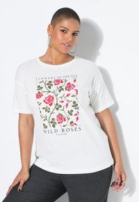 Personne aux cheveux courts portant des boucles d'oreilles créoles et un t-shirt blanc avec un imprimé floral rose et vert et le texte "Roses Sauvages", posant contre un mur blanc.