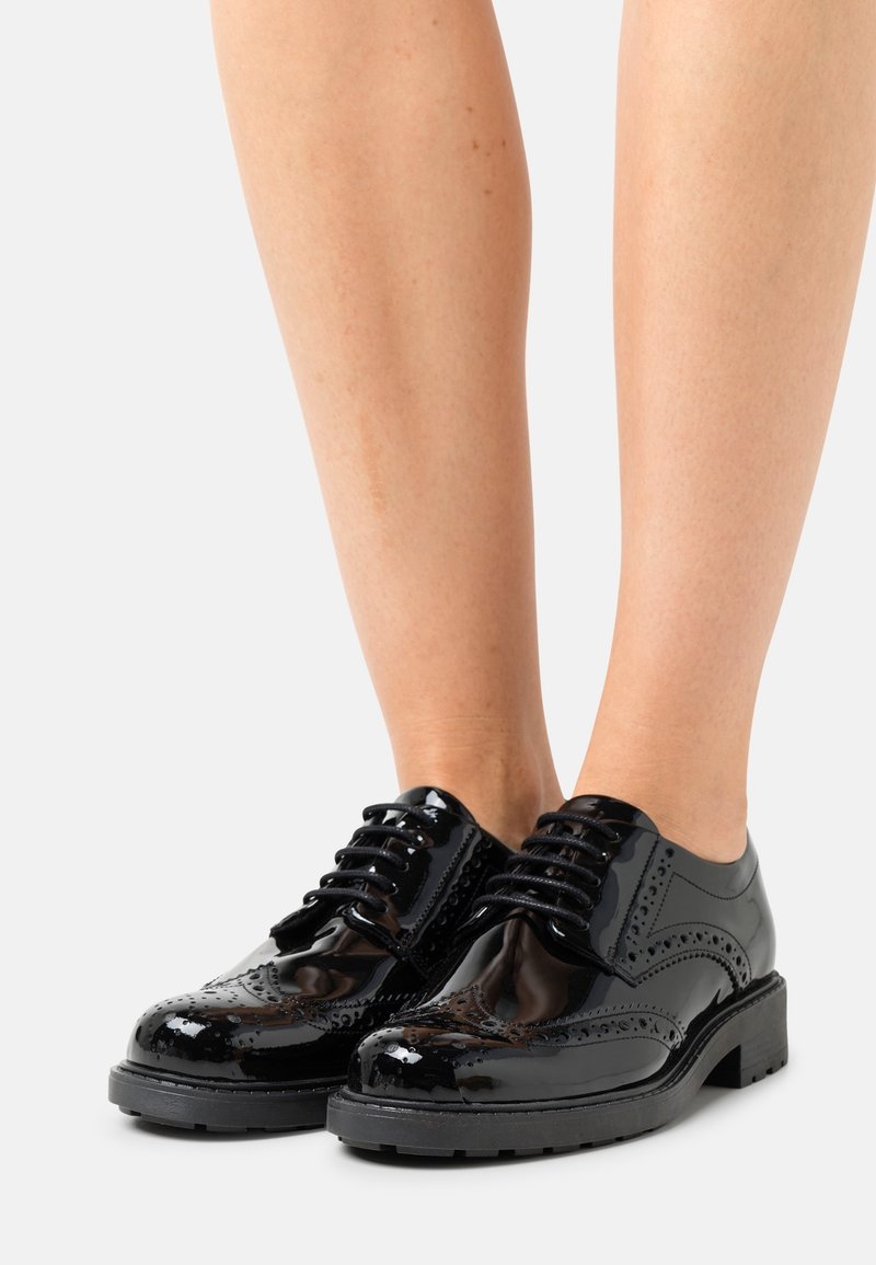 Chaussures brogue en cuir verni noir brillant avec des semelles épaisses et des lacets portées sur des jambes nues sur un fond uni.