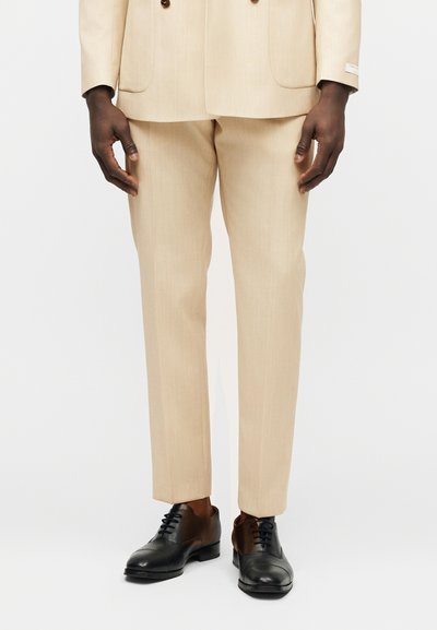 Άνδρας ντυμένος με μπεζ παντελόνι κοστούμι ραμμένο στα μέτρα και ταιριαστό σακάκι, φορώντας μαύρα δερμάτινα παπούτσια επίσημου στυλ, στέκεται μπροστά σε λευκό φόντο.