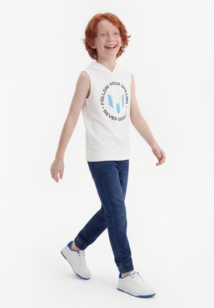 Παιδί με κόκκινα μαλλιά που περπατάει και χαμογελάει, φοράει αμάνικο λευκό φούτερ με το κείμενο «Ακολούθησε τα όνειρά σου, μην τα παρατάς ποτέ», μπλε τζιν και λευκά αθλητικά παπούτσια.