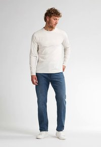 Cămașă bej deschis cu mâneci lungi, asortată cu blugi albaștri din denim și adidași albi. Cămașa are un decolteu rotund și un croi mulat.