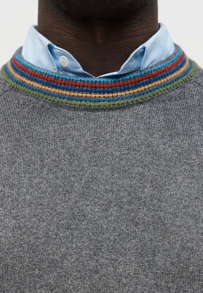 Personne à la peau foncée portant une chemise bleu clair boutonnée sous un pull gris avec un col rayé multicolore.