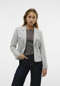 Blazer gris ajustado con una textura suave y solapas de muesca, combinado con una blusa oscura y jeans. El diseño cuenta con un cierre de un solo botón.
