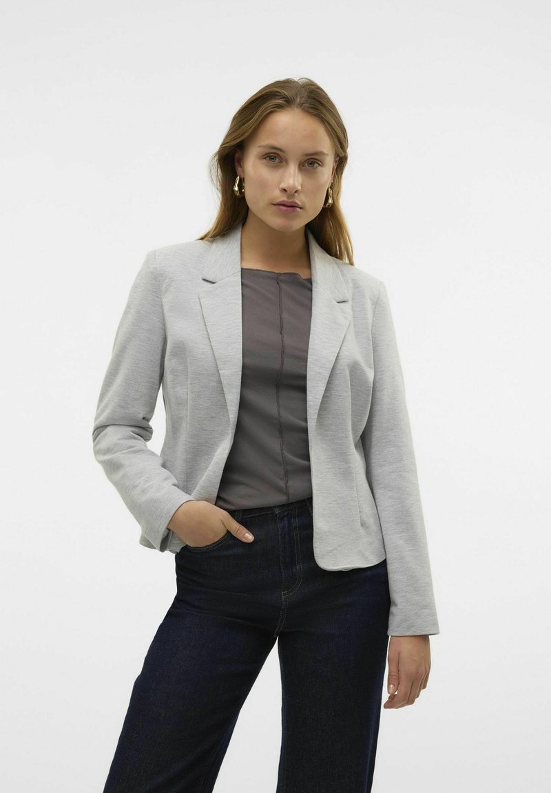 Blazer gris ajustado con una textura suave y solapas de muesca, combinado con una blusa oscura y jeans. El diseño cuenta con un cierre de un solo botón.