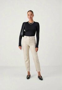 Black long-sleeve top, beige high-waisted trousers, and black flat shoes. Model wears a chain necklace and carries a black bag.
