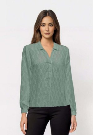 Blusa verde chiaro a maniche lunghe con tessuto testurizzato a motivo diamante, dotata di colletto e chiusura a bottoni. Indossata con pantaloni neri.