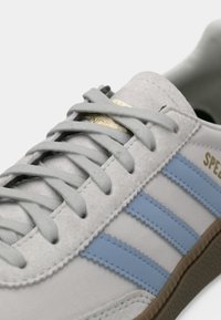 Sapatilha cinza em camurça com riscas azul claro, sola texturizada e atacadores cinzentos planos. Detalhe do logótipo dourado na lateral. Forma do bico arredondada.