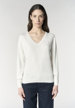 Donna con lunghi capelli castani che indossa un maglione bianco a scollo a V e pantaloni scuri, in piedi davanti a uno sfondo chiaro e uniforme.