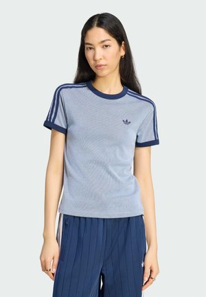 Kvinde med langt mørkt hår iført en lysegrå Adidas T-shirt med navy striber og navy blå plisserede bukser med sidelommer med lynlås.