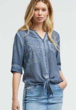 Woman wearing a blue button-up shirt with white text graphic tied at the waist, paired with a light blue denim skirt, against a plain background.