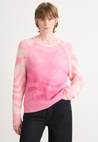 Vrouw met kort bruin haar, gekleed in een roze geblokte trui en zwarte broek, staand met één hand in de zak tegen een effen achtergrond.