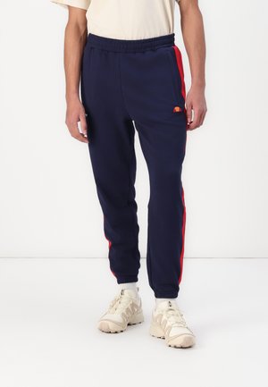 Pantalones de chándal azul marino con franjas laterales rojas, cinturilla elástica y tobillos ajustados. Hechos de un tejido suave, con un pequeño logotipo en el muslo.
