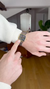 Ceas cu ramă aurie și cadran negru care afișează modele colorate și abstracte. Are o curea din piele maro. O mână indică ceasul.