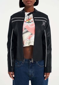 Svart läderjacka med vita randiga detaljer, dragkedjestängning och knappkrage. Bärs över en grafisk crop top med blå denimjeans.