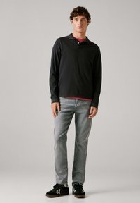 Hombre joven de pie vistiendo una camiseta de manga larga negra tipo polo, camiseta interior roja, vaqueros gris claro y zapatillas negras sobre un fondo gris liso.